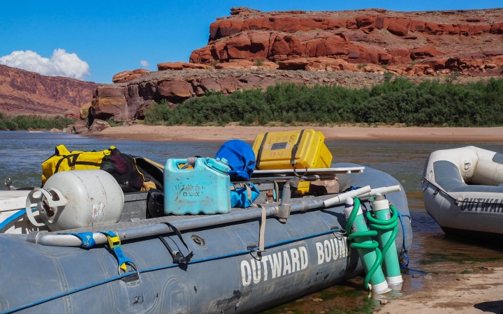 A beached raft rests on the shore of a river. There are red canyon walls in the background. 